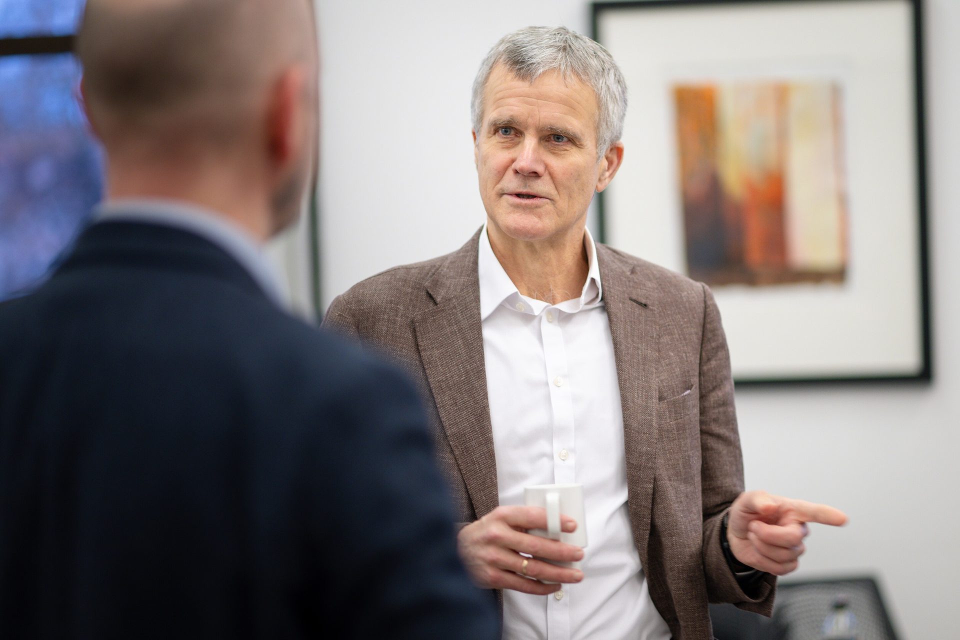 Middle-aged man in brown jacket talks, holds mug, points to person with back to camera.