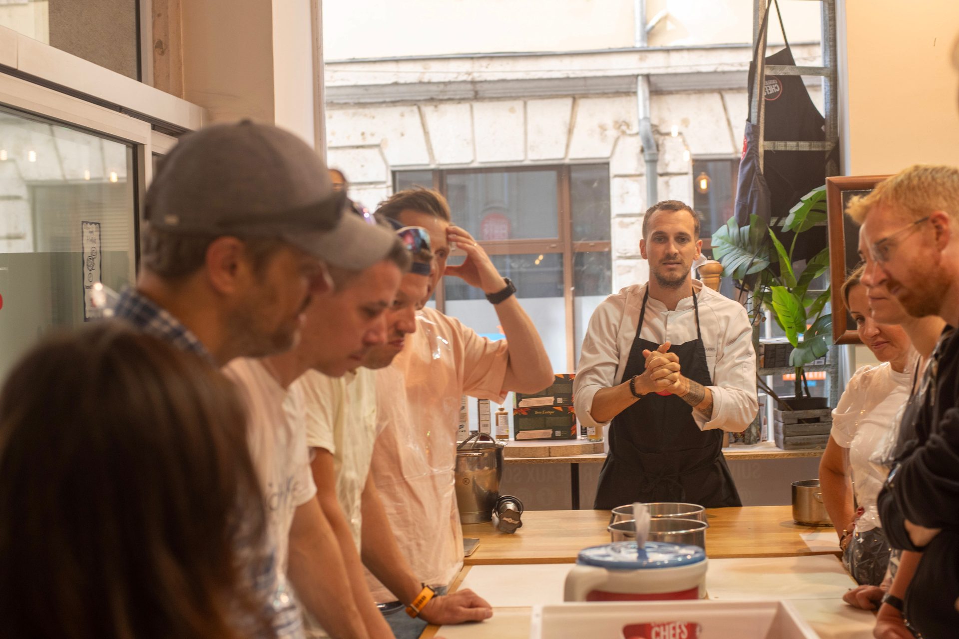 A chef in a black apron instructs a group of people gathered at a cooking class counter.