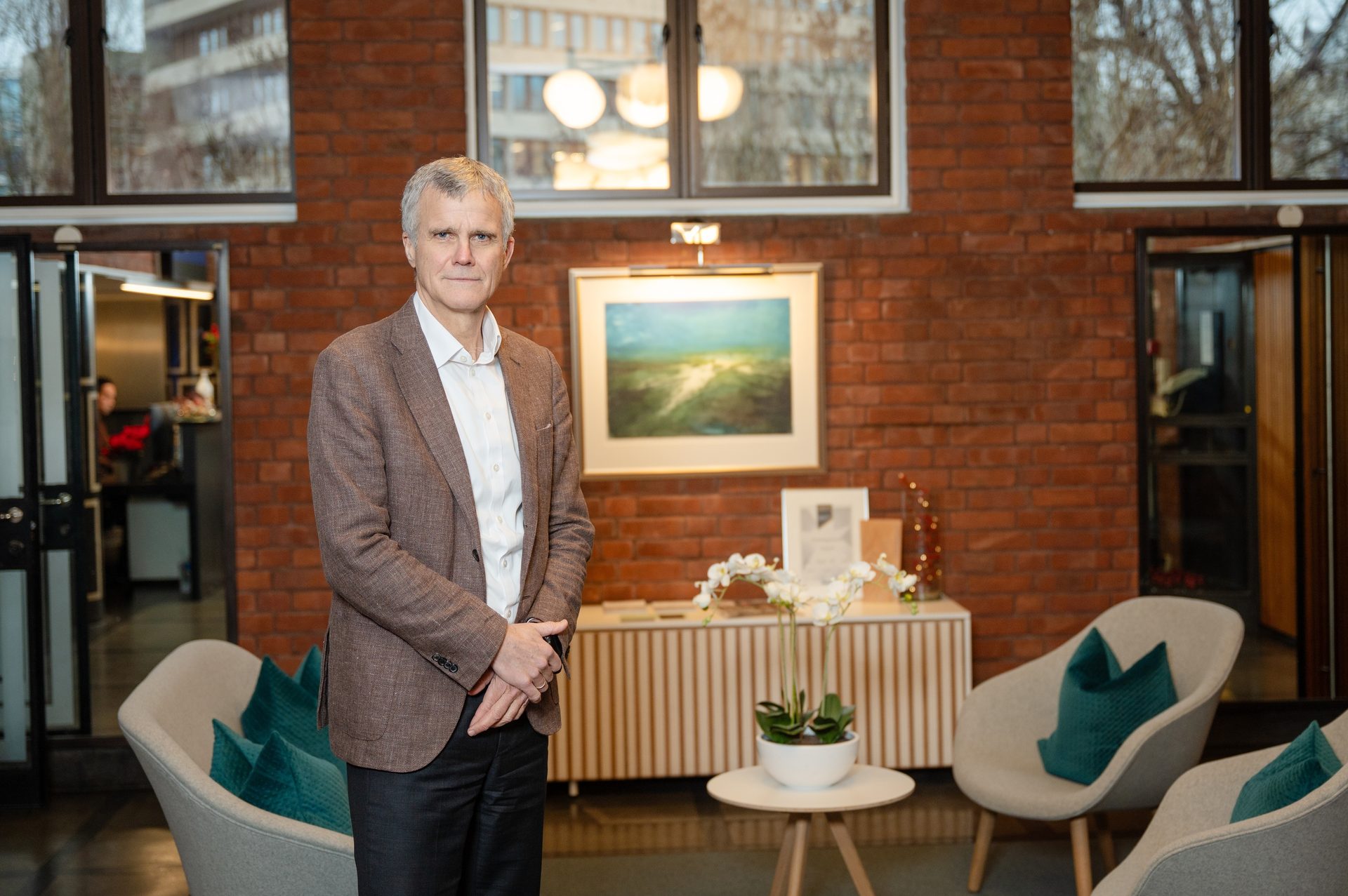 A man in a brown jacket stands in a modern lobby with brick walls, windows, and stylish chairs.