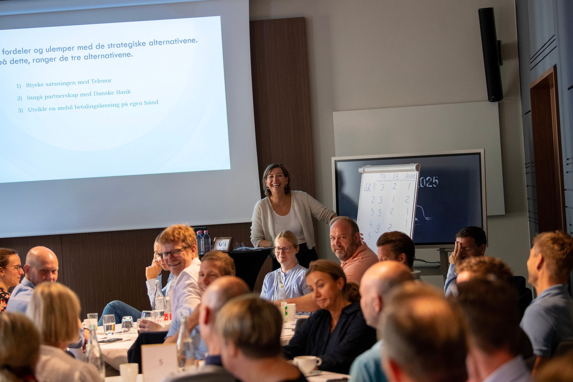 A woman smiles while presenting to a room full of people, with a strategic alternatives slide visible.