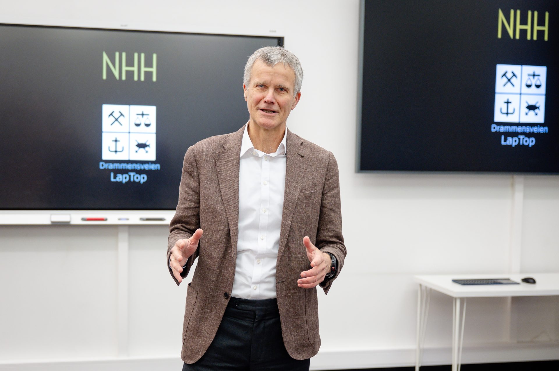 Man in brown jacket gesturing in front of screens with 'NHH' logo.