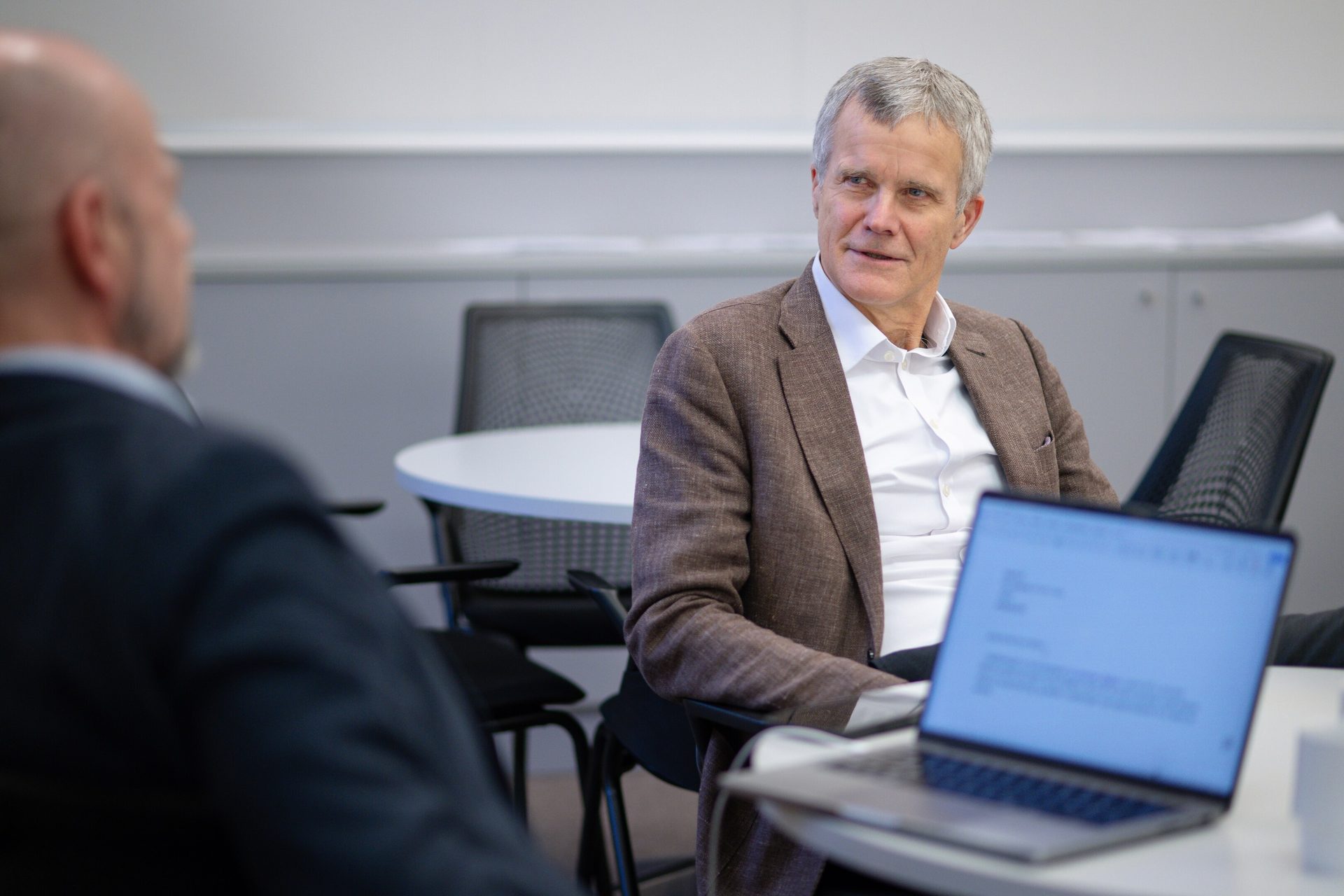Man in brown jacket listening to another man in an office. Laptop on table.