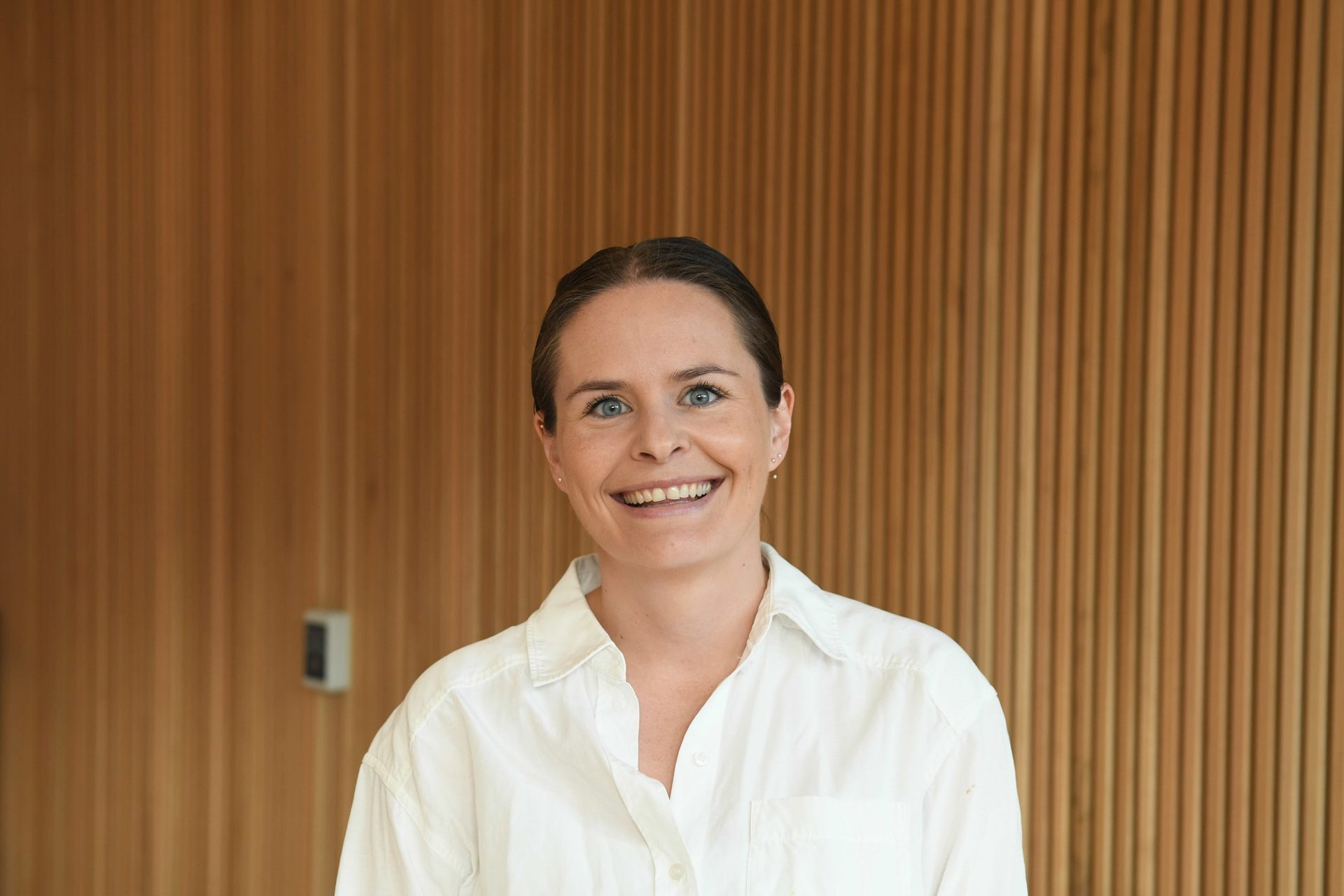 A smiling woman with blue eyes and brown hair, wearing a white shirt, against a wooden slatted wall.