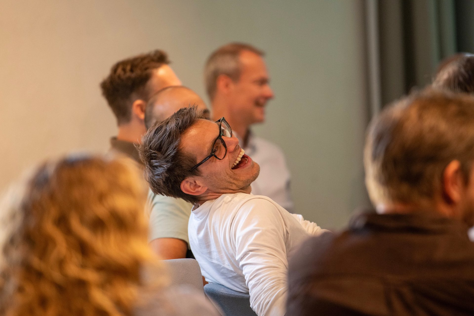 Man with glasses laughing heartily, head thrown back, in a group setting.