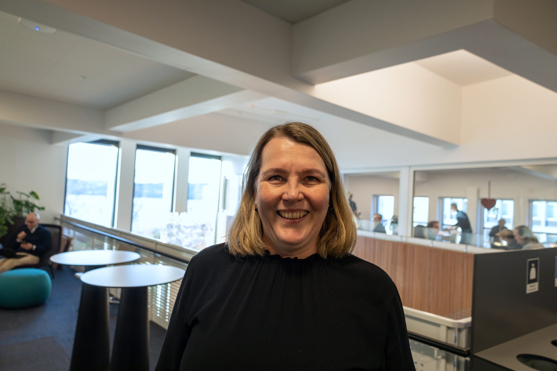 A smiling woman with blonde hair, dressed in black, stands indoors with a blurry background.