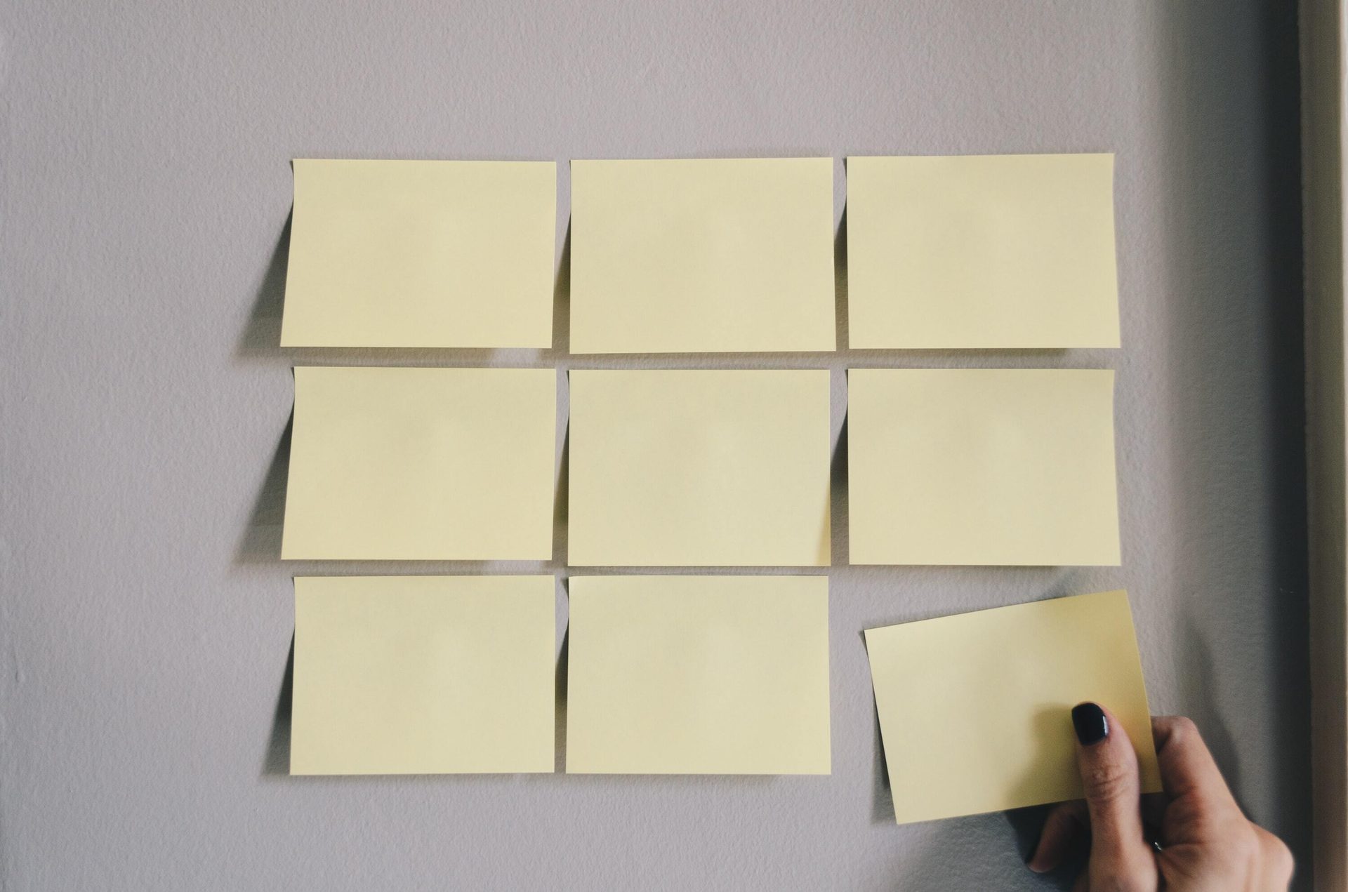 Blank yellow sticky notes arranged on a gray wall, with a hand holding one.