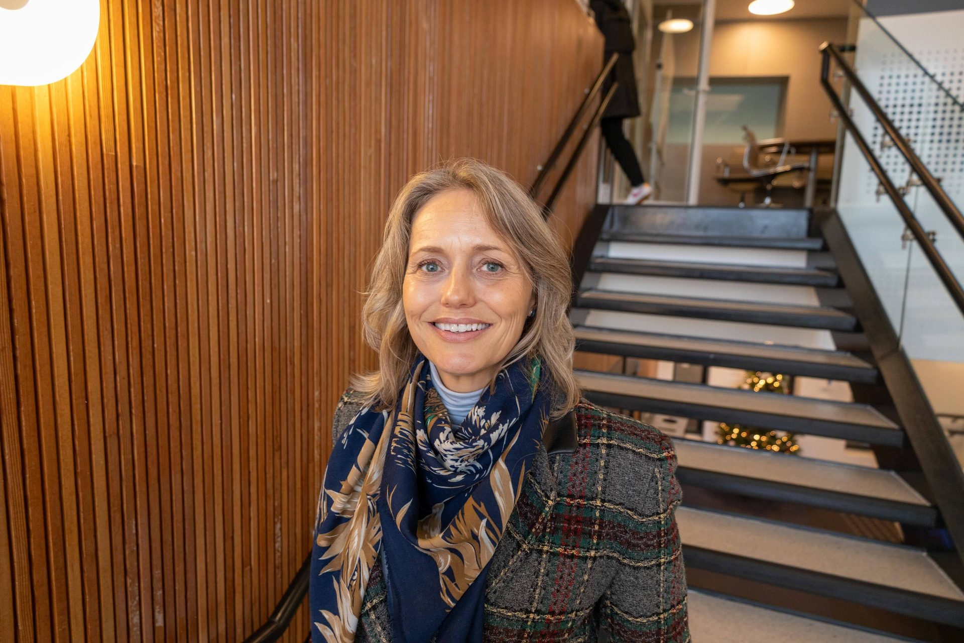 Woman in plaid jacket and scarf smiling in an office setting with wooden walls and stairs.