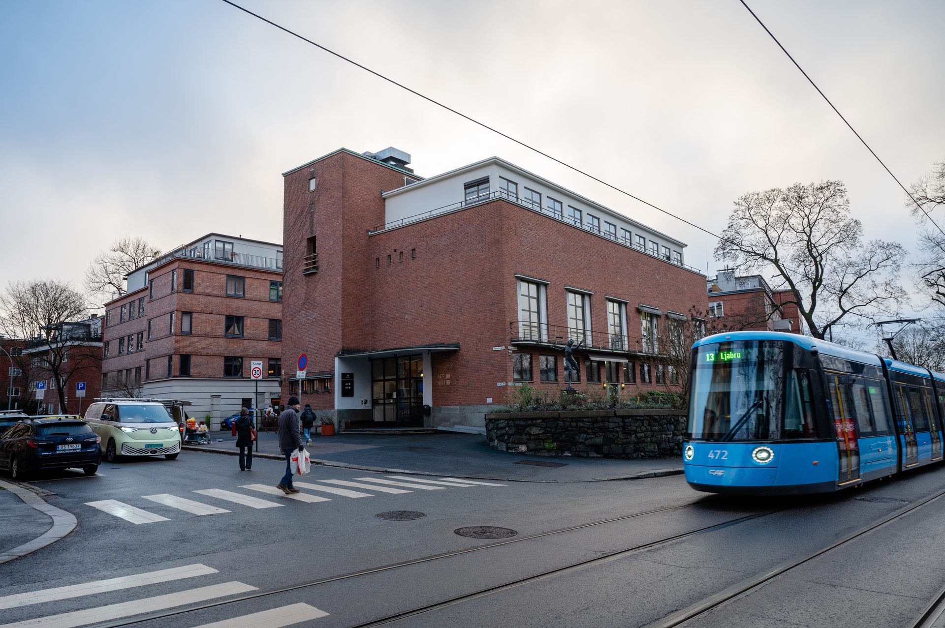 A blue tram on tracks, people on a crosswalk, and a modern brick building in a city street.