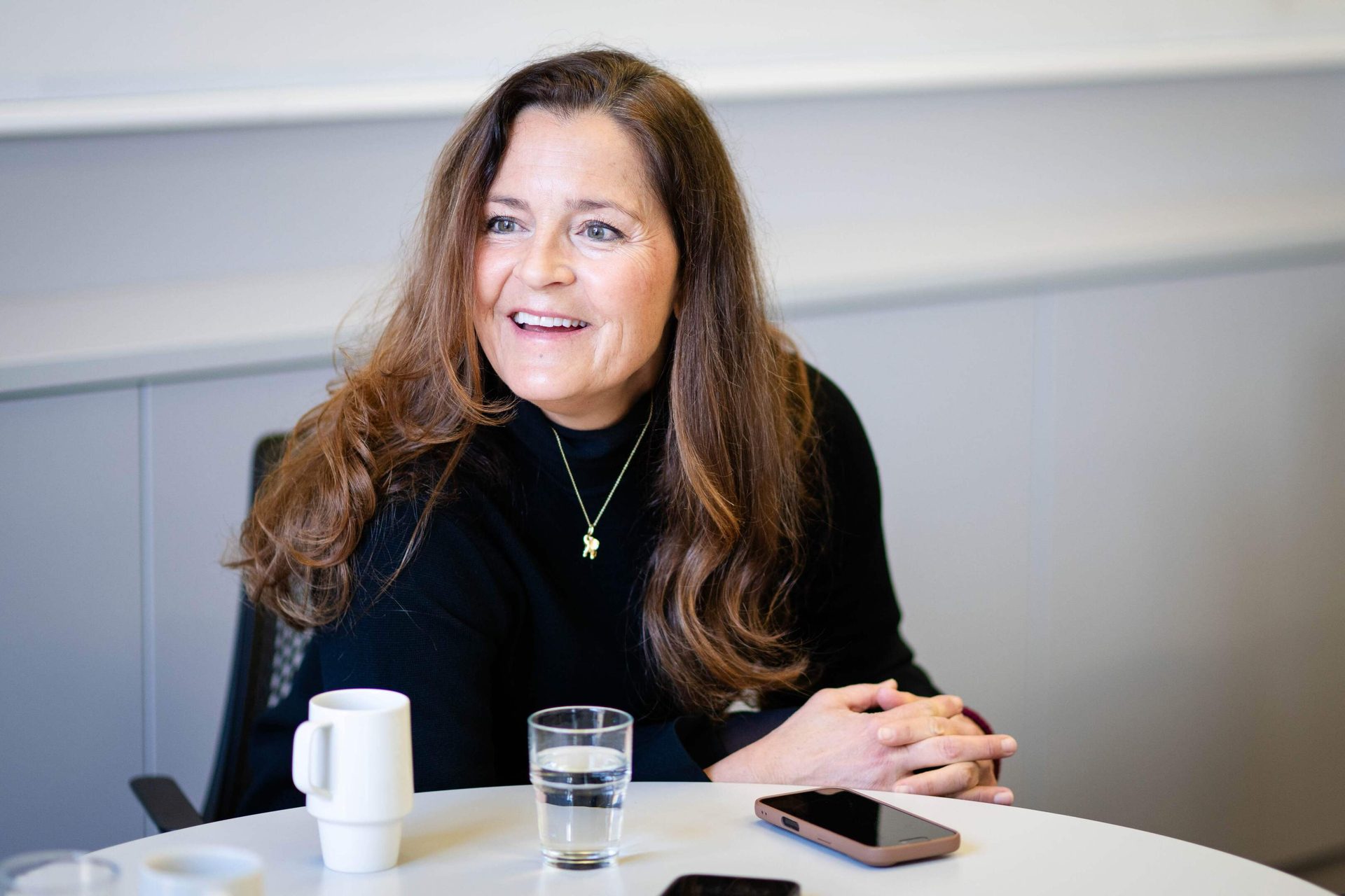 Smiling woman with long brown hair, black top, elephant necklace, sitting at a table.