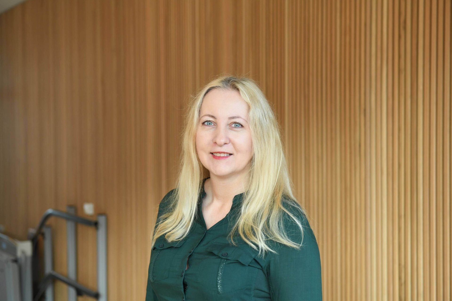 A smiling woman with blonde hair and blue eyes wears a green shirt in front of a wooden wall.