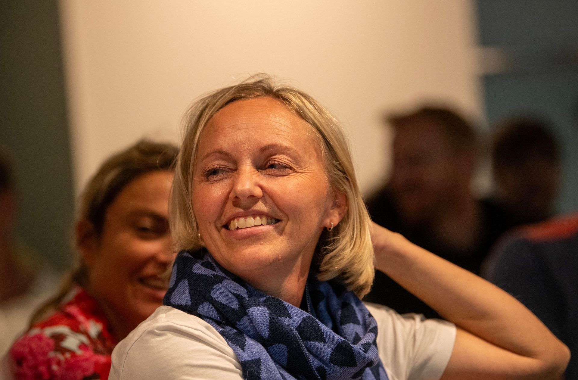 Smiling blonde woman in a white shirt and blue patterned scarf, with her hand behind her head.
