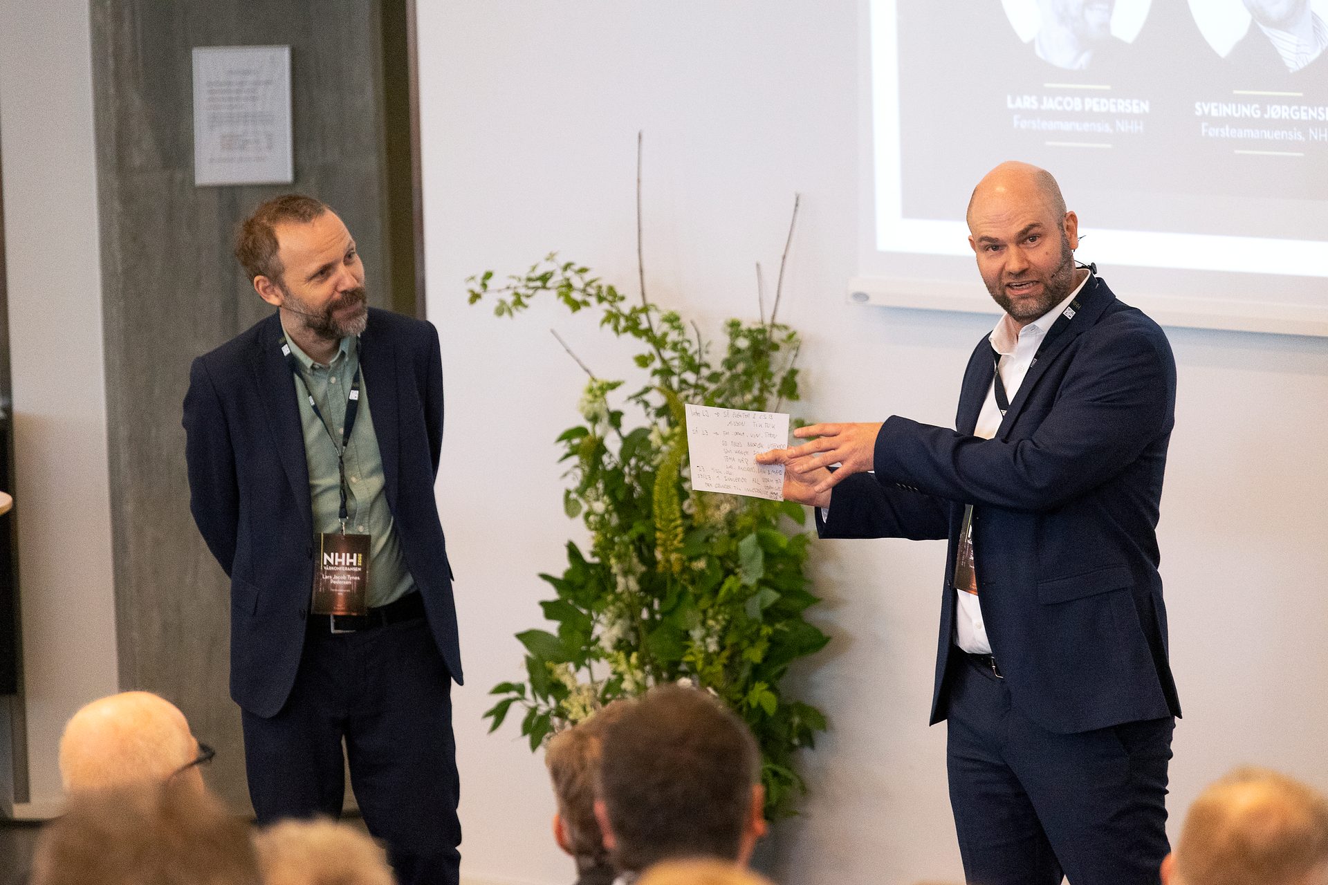 Two men in suits at a conference, one presenting with a note, the other listening attentively.