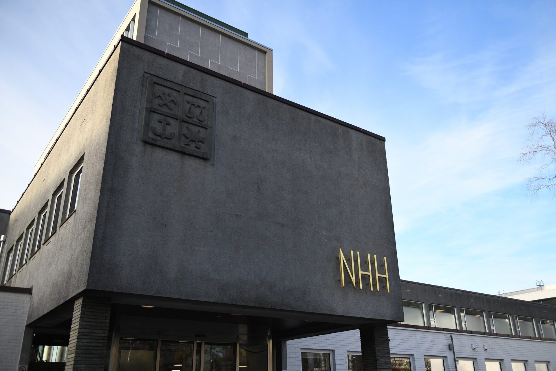 Grey NHH building with gold letters and a crest, under a blue sky.
