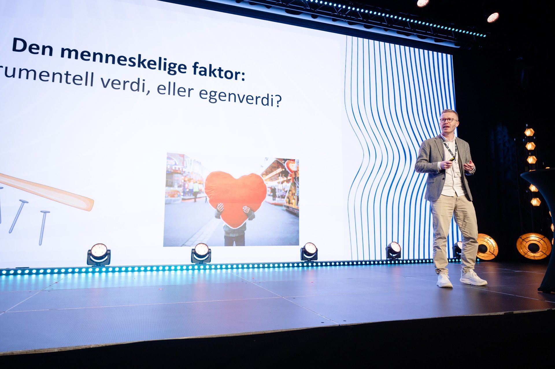 A man presents on stage with a large screen behind him displaying the text "The human factor" and an image of a person holding a heart.