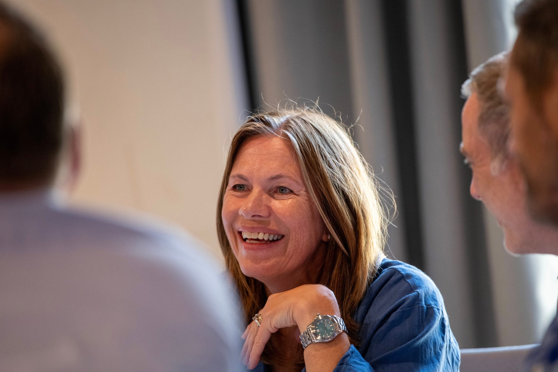 A smiling woman with long brown hair laughs, looking towards others at a table.