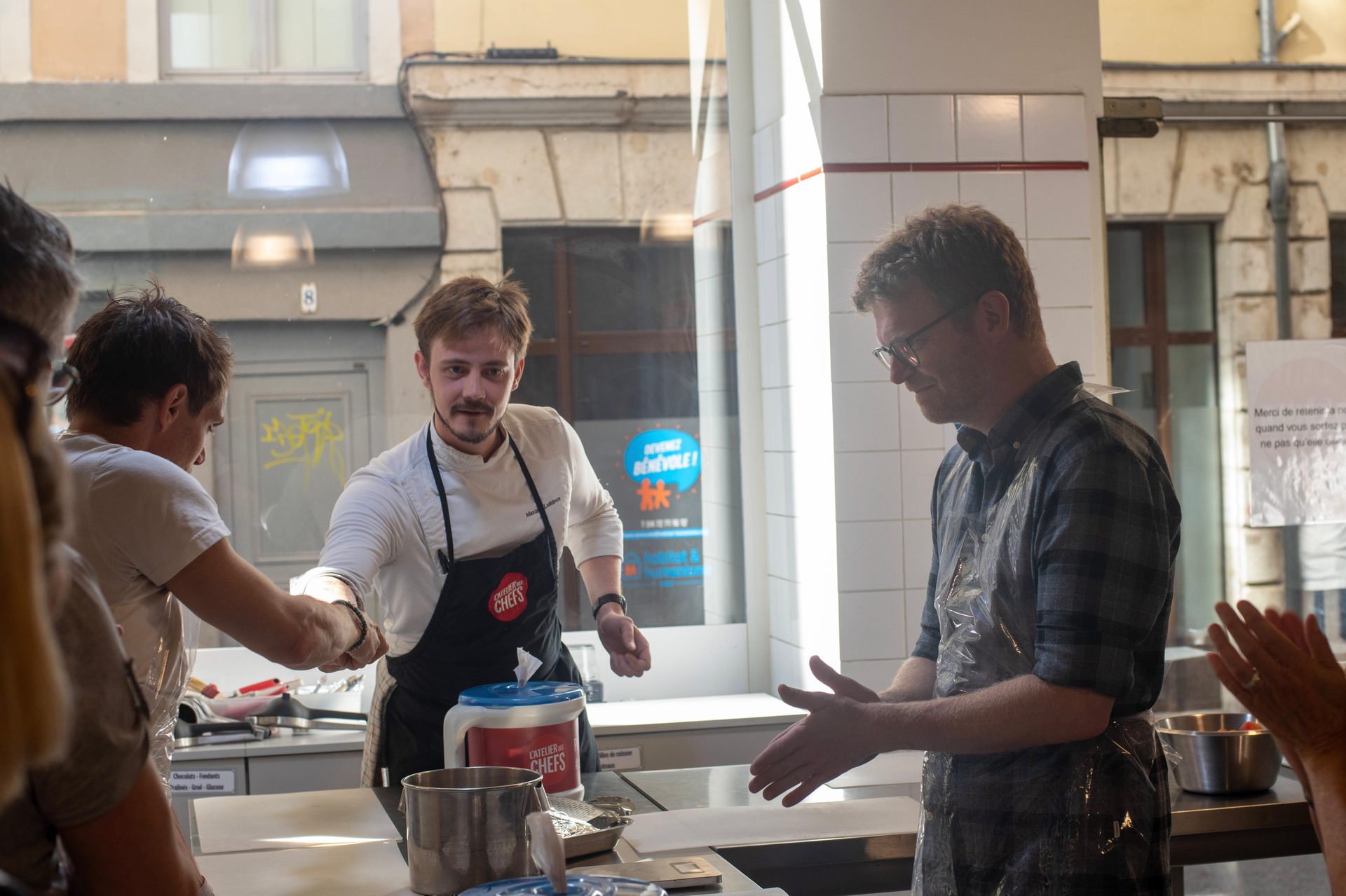 Two chefs interact in a kitchen or cooking class; one shakes hands, another gestures.