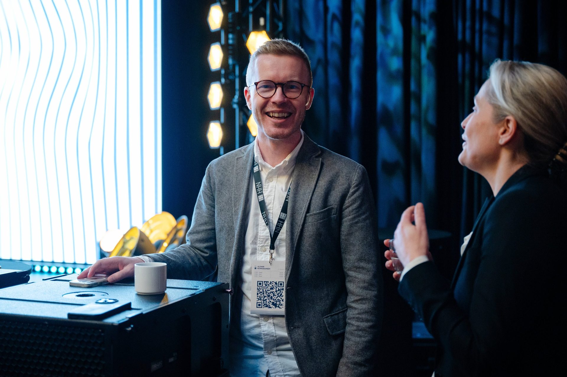 Two people, a man in glasses and a woman, engaged in conversation at an event with stage lighting.