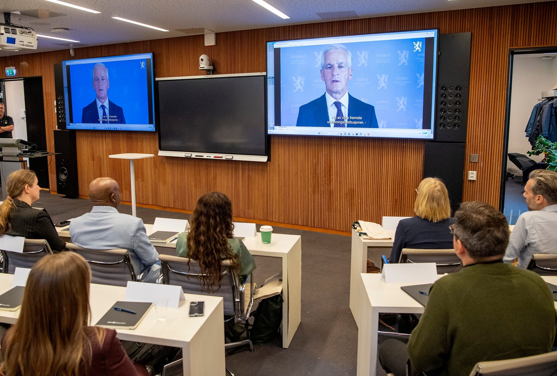 People in a conference room watching a man speak on two large video screens.