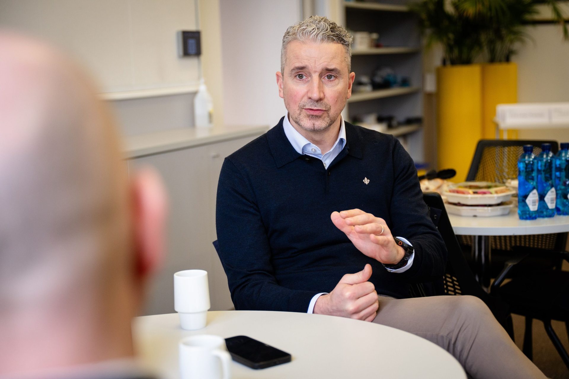 A man with grey hair and a navy sweater speaks, gesturing with his hands across a table to another person.