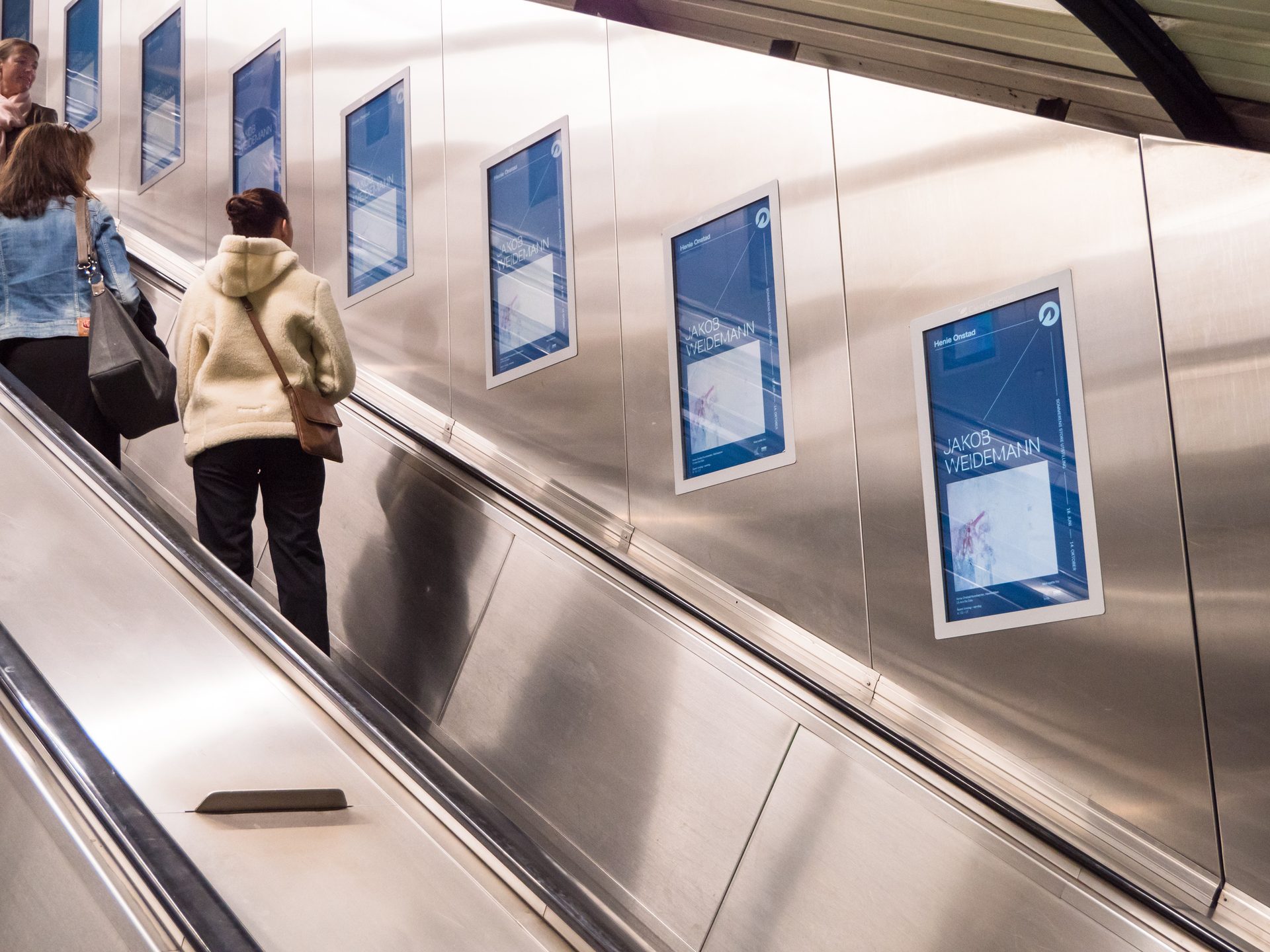 Women on an escalator next to multiple digital screens displaying "Jakob Weidemann" exhibition posters.