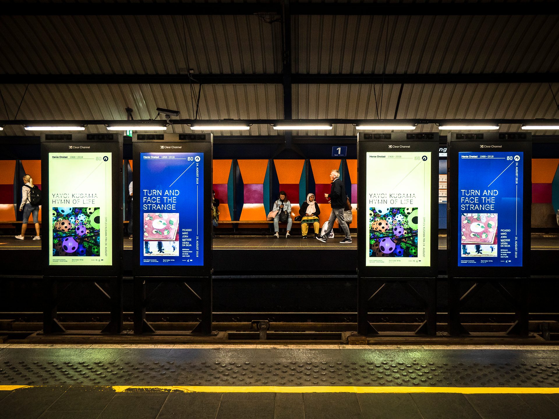 People wait and walk on a subway platform next to four brightly lit digital advertisement boards.