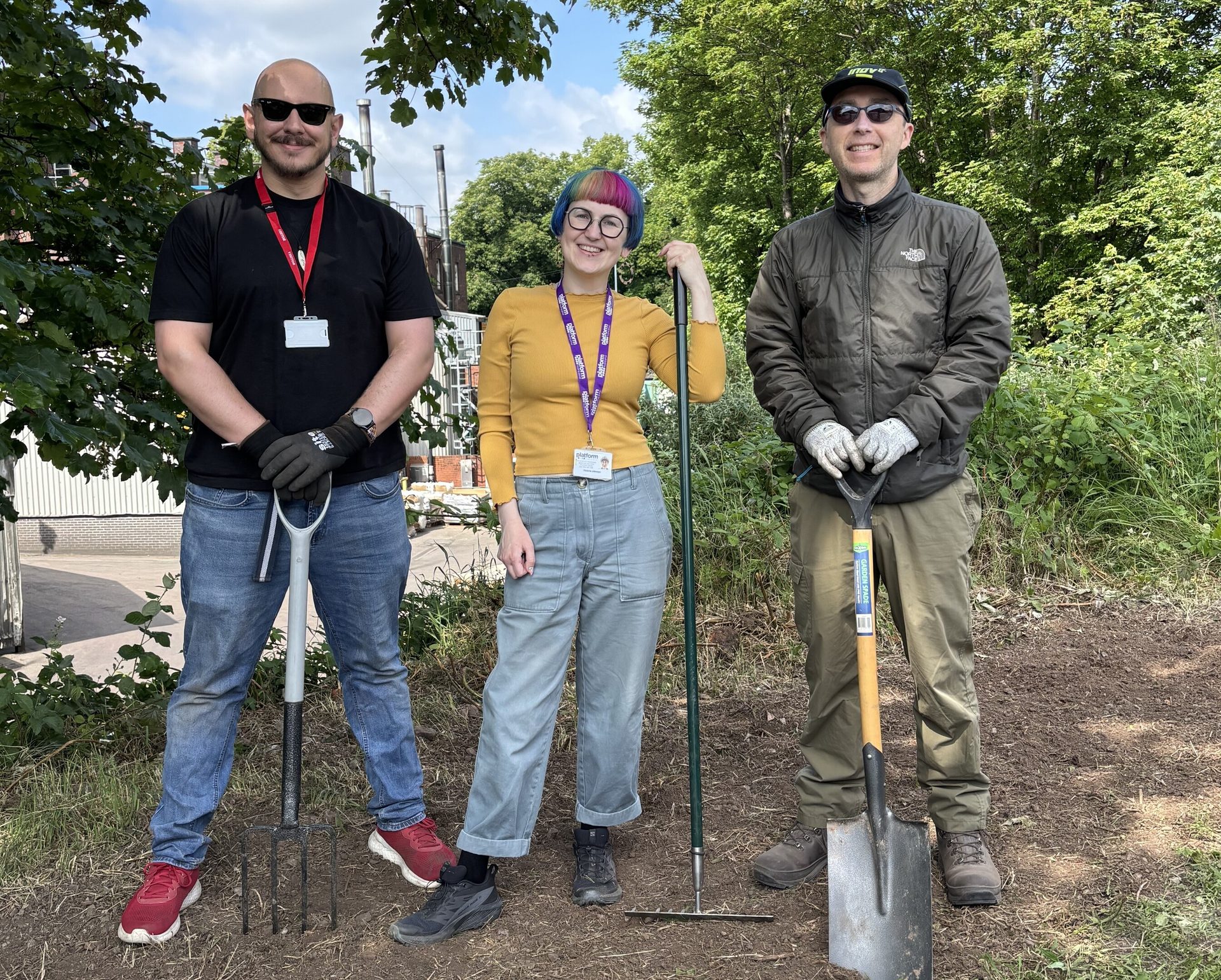 Three smiling people holding a pitchfork, rake, and shovel outdoors.