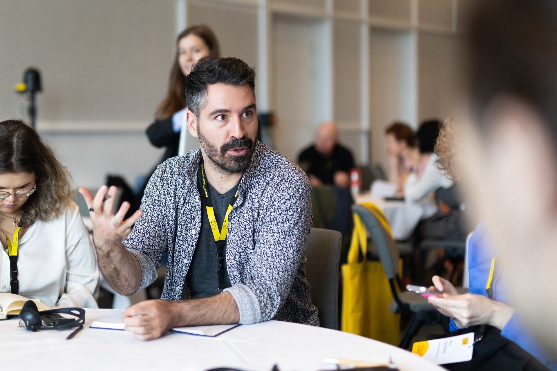 A man with a beard gestures while speaking at a conference table, with other attendees in the foreground and background.