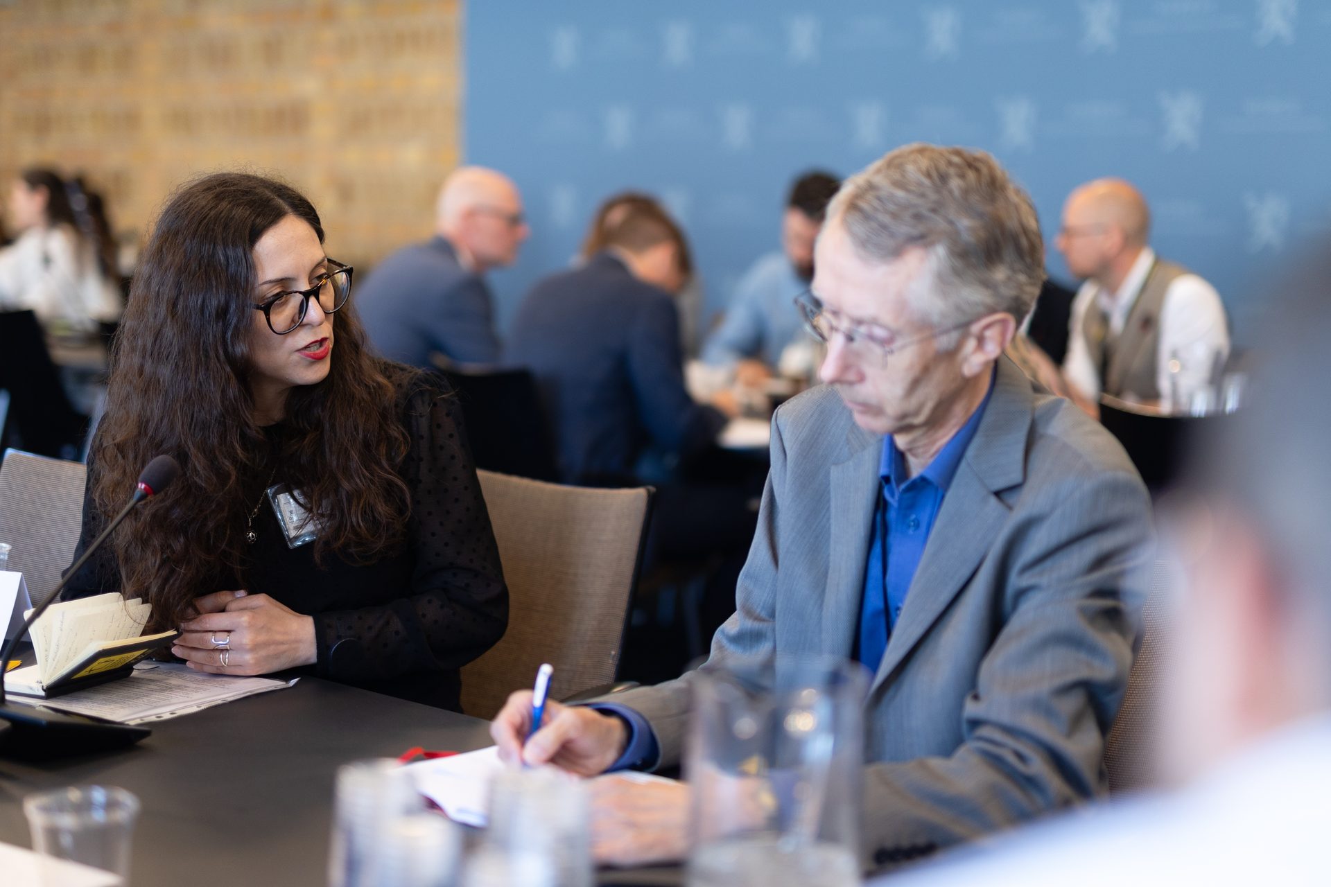 A woman speaking into a microphone and a man taking notes at a conference table during a meeting.