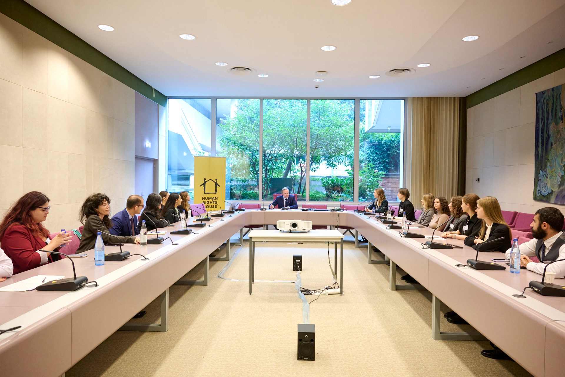 U-shaped meeting with attendees, a speaker, and a 'Human Rights House' banner.