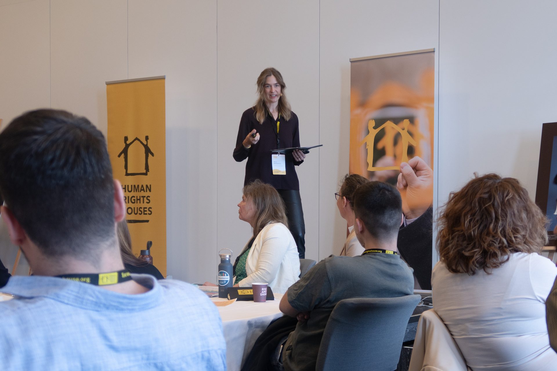 A woman presents to an audience at an event featuring "Human Rights Houses" banners.