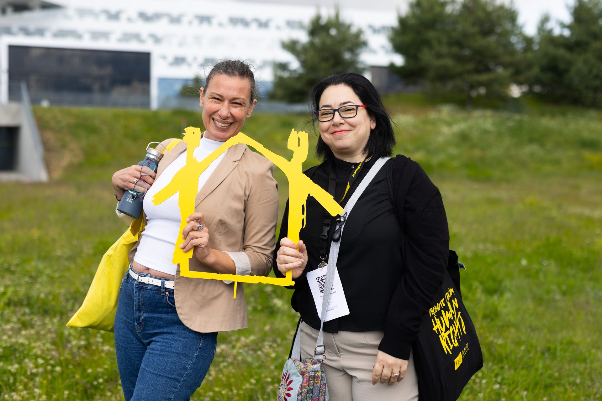 Two smiling women holding a yellow, house-shaped frame made of stick figures against a grassy background.