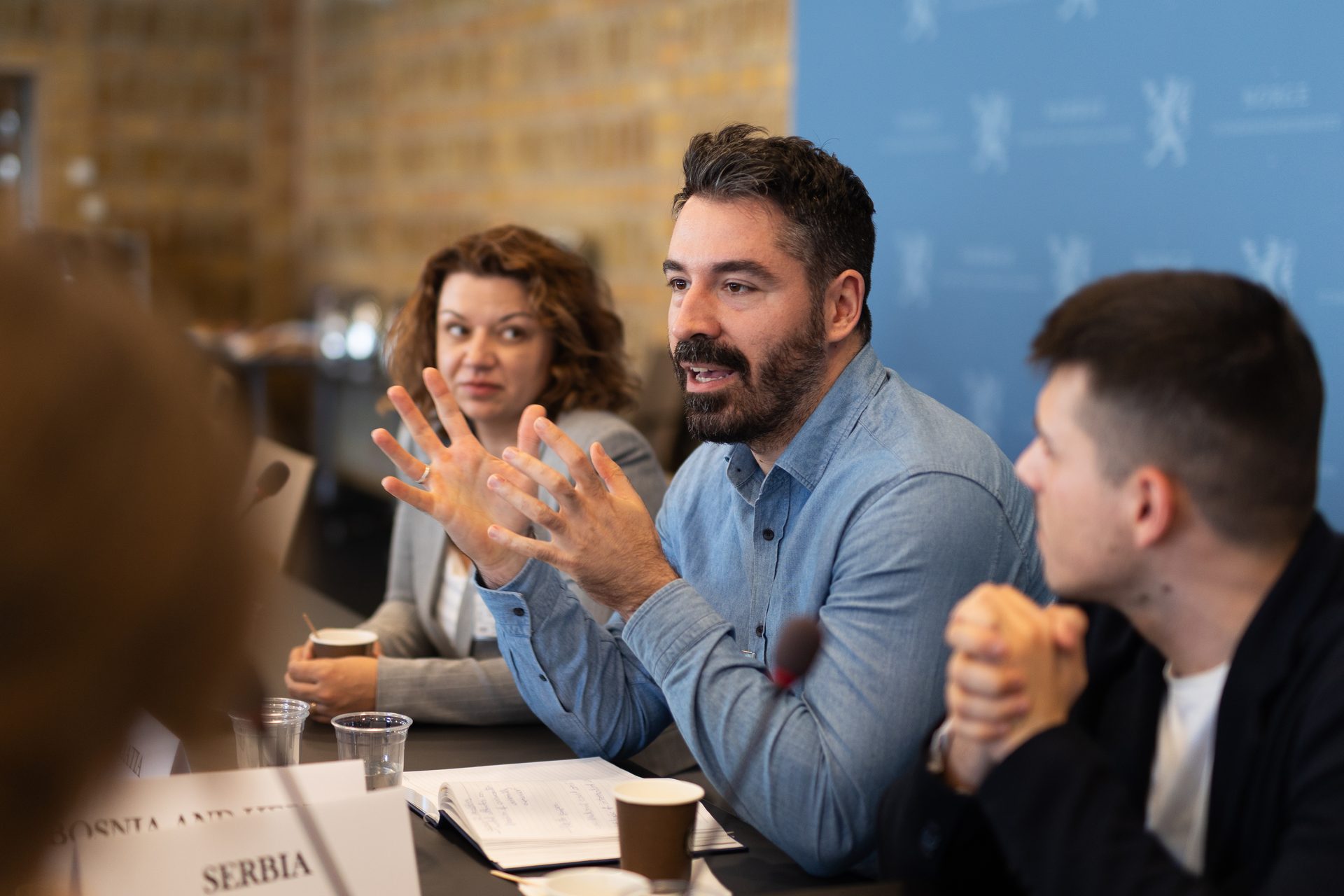 Man speaking and gesturing at a meeting table with two other attendees.