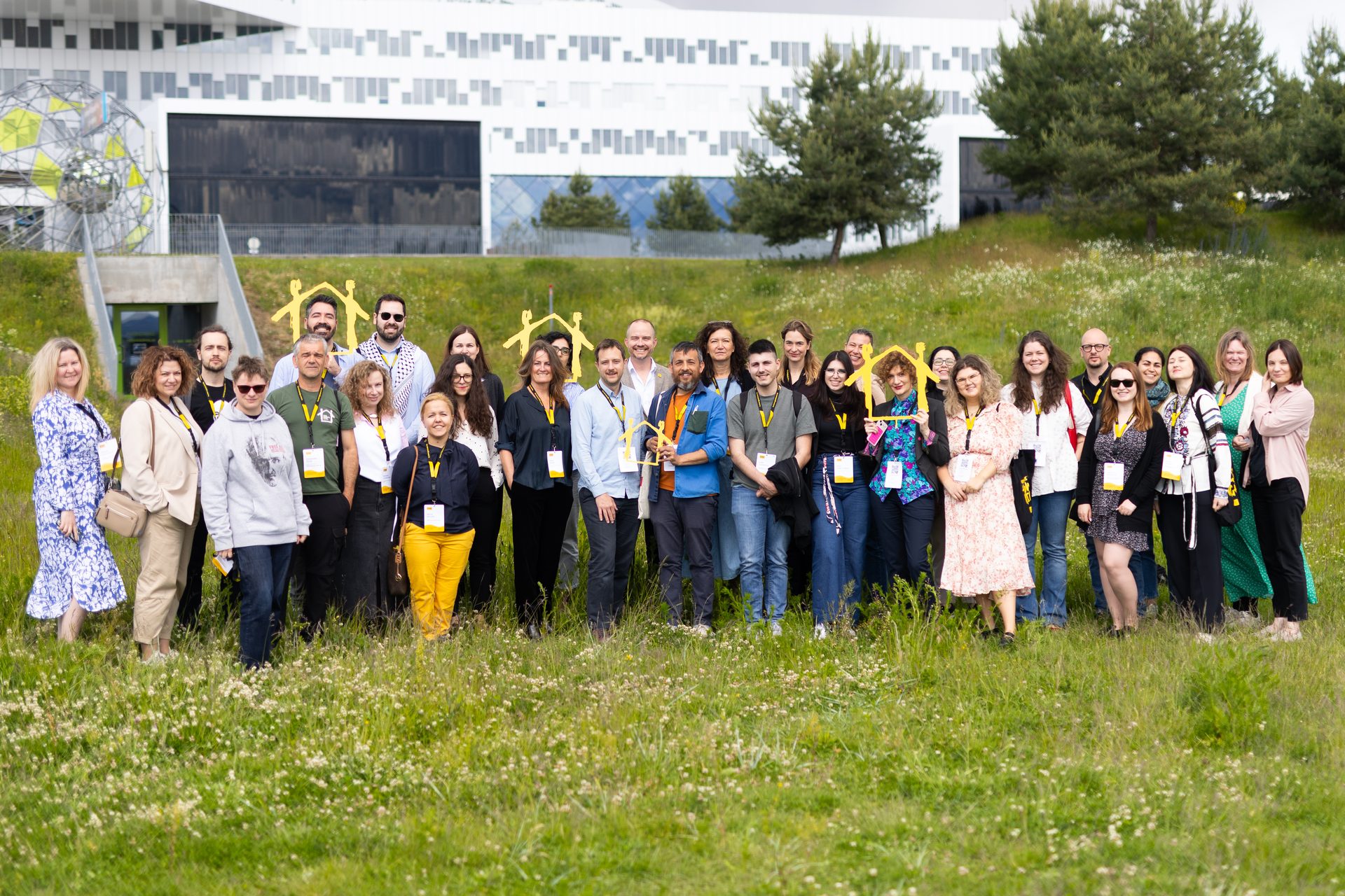 A large group of diverse people posing outdoors in a grassy field with a modern building behind them.