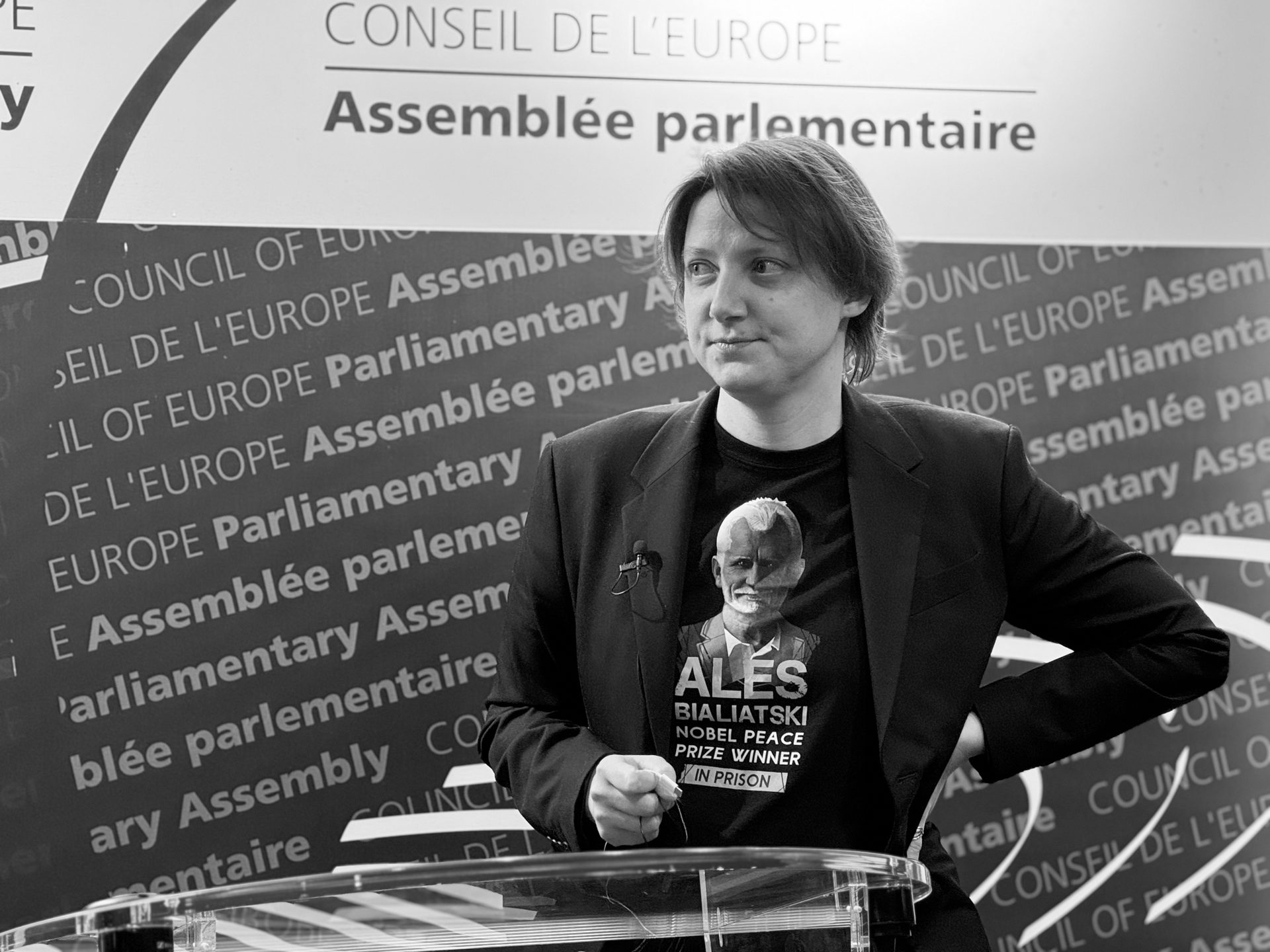 A woman stands at a podium, wearing a t-shirt featuring Ales Bialiatski, Nobel Peace Prize winner in prison.