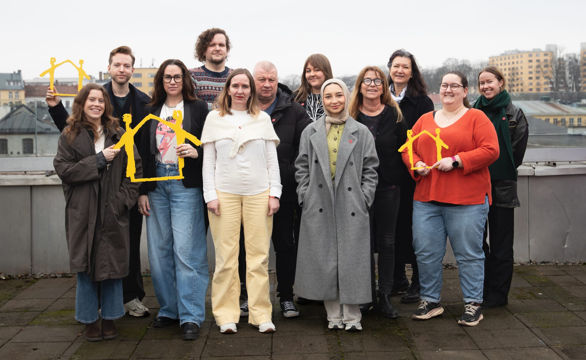 A diverse group of people, men and women of various ages, stand together holding yellow house shapes.
