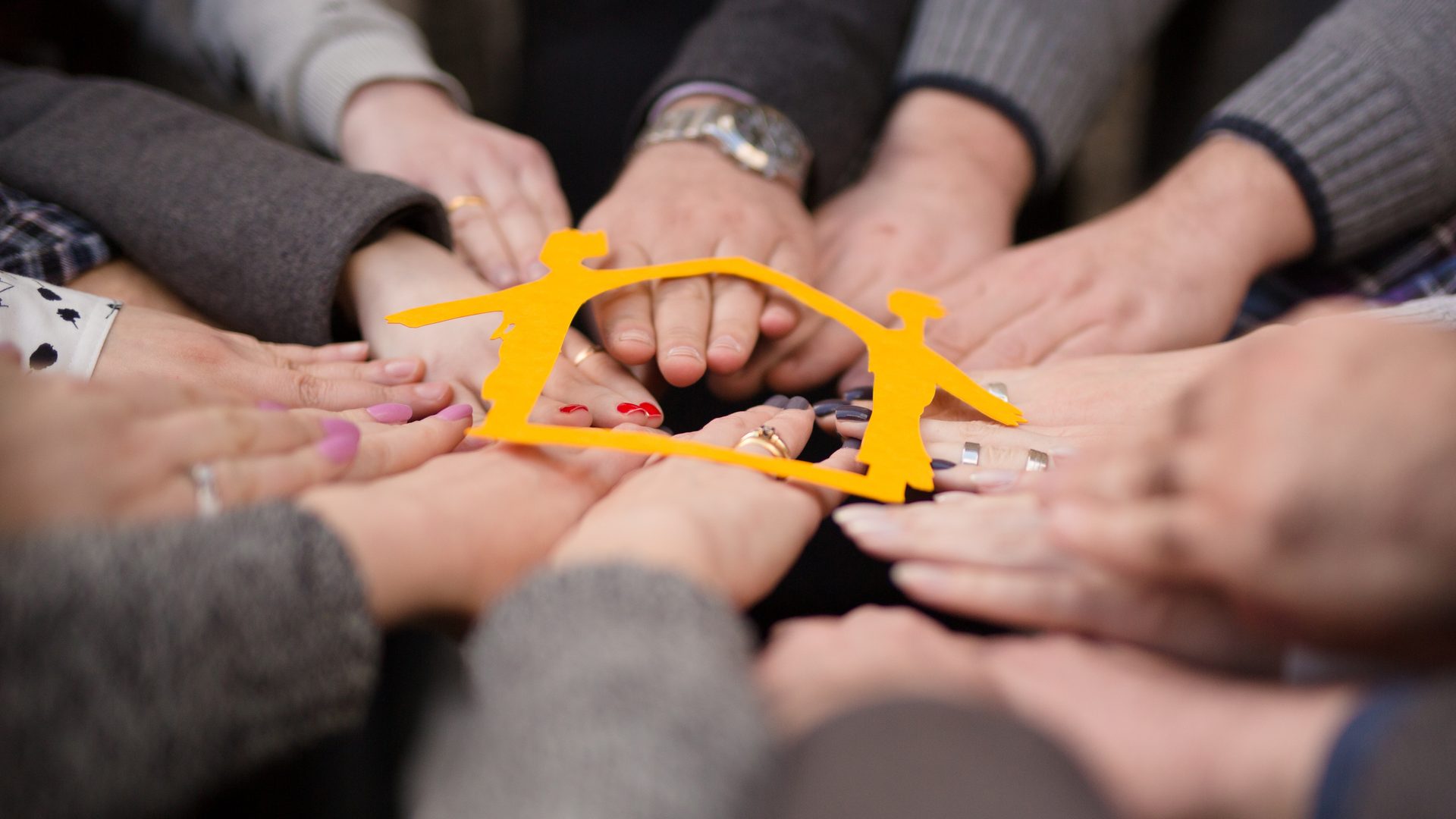 Diverse hands come together in a circle, holding an orange cutout of people forming a house.
