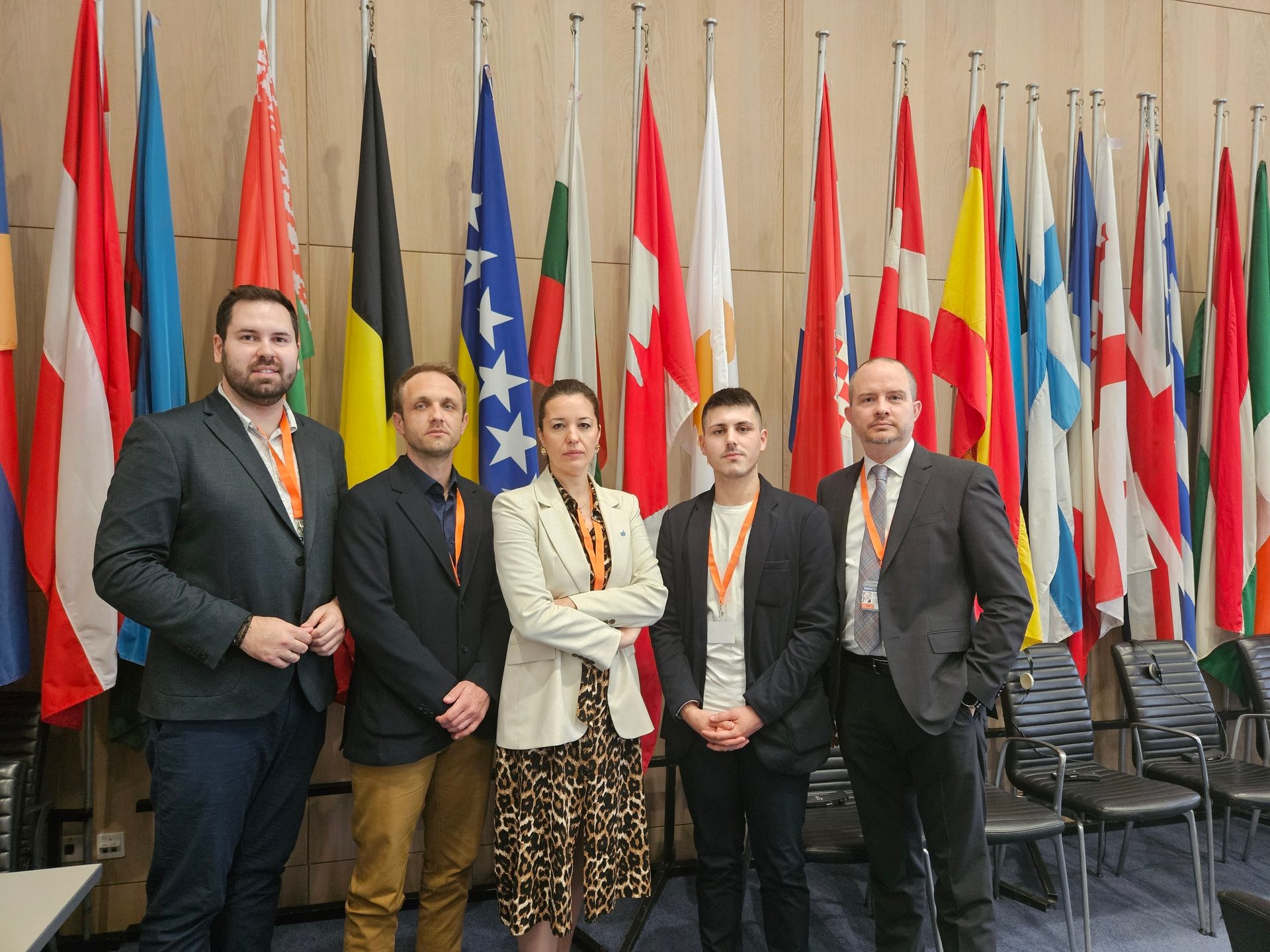 Five professionals (four men, one woman) stand in front of multiple national flags.