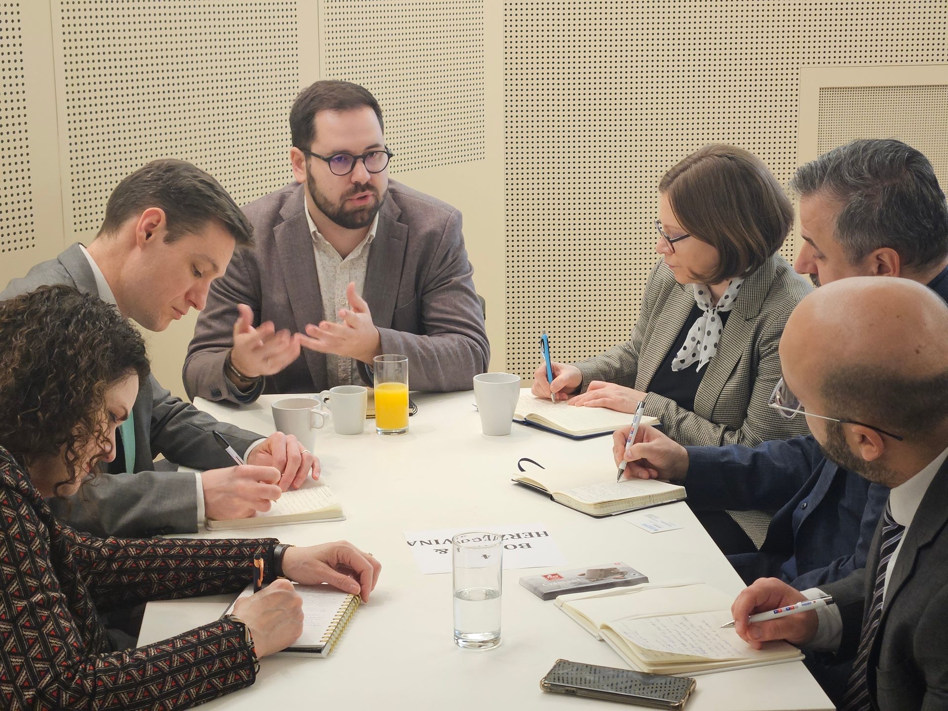 Six people, four taking notes, and one speaking, seated around a table during a meeting with drinks.