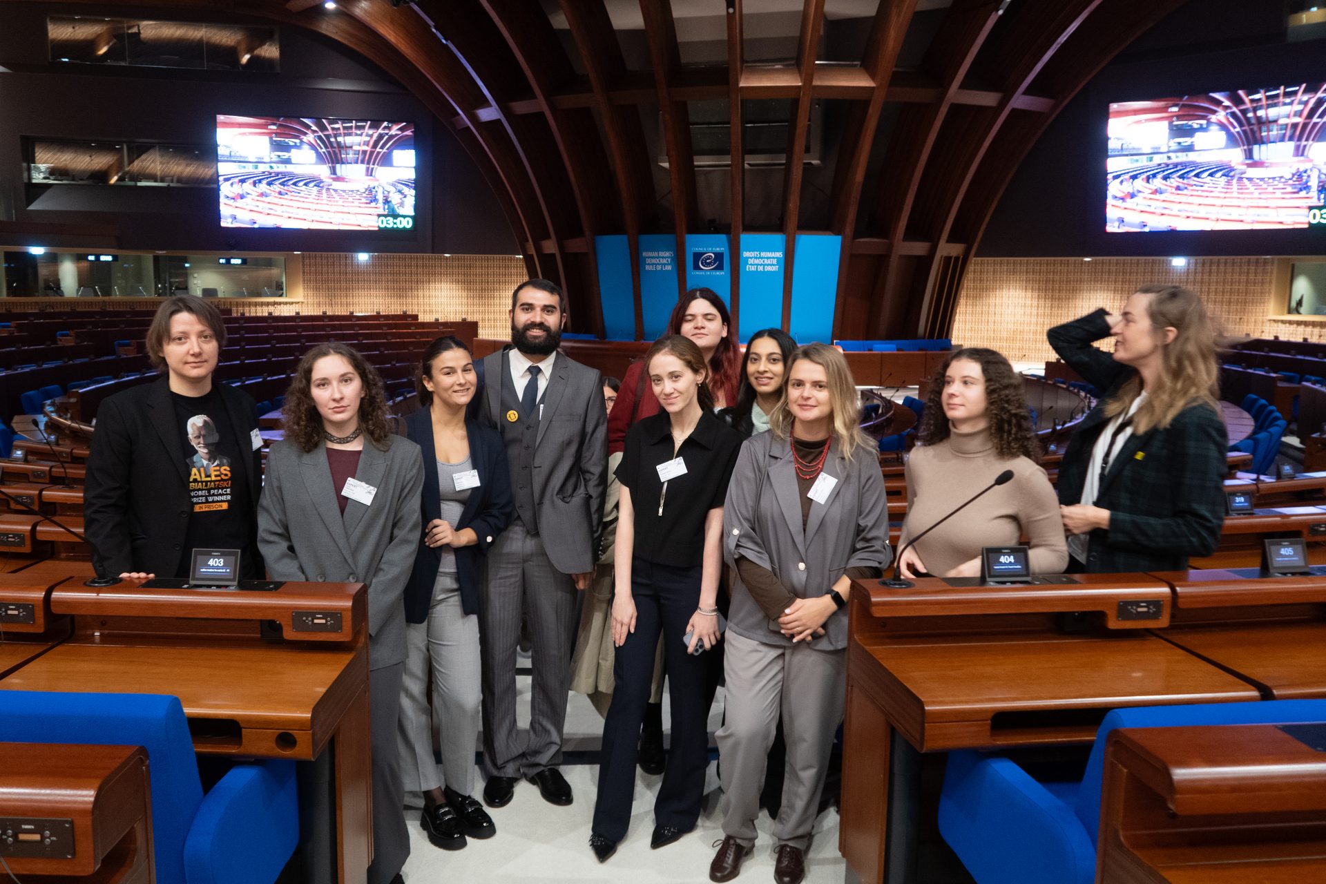 A group of ten young people pose in an assembly hall with wooden desks and blue chairs.