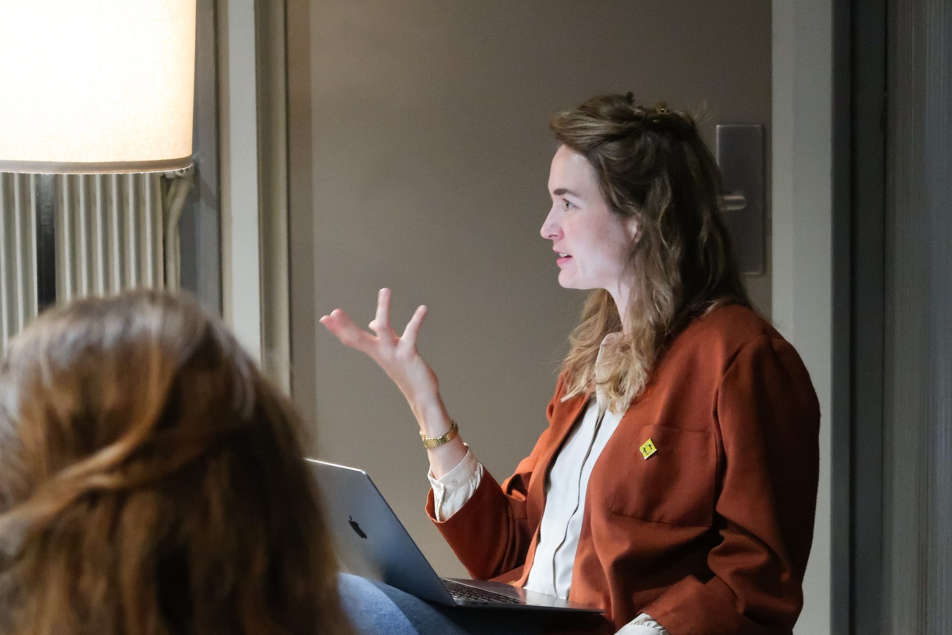 A woman with long hair wearing an orange jacket speaks, gesturing with her hand, a laptop on her lap.