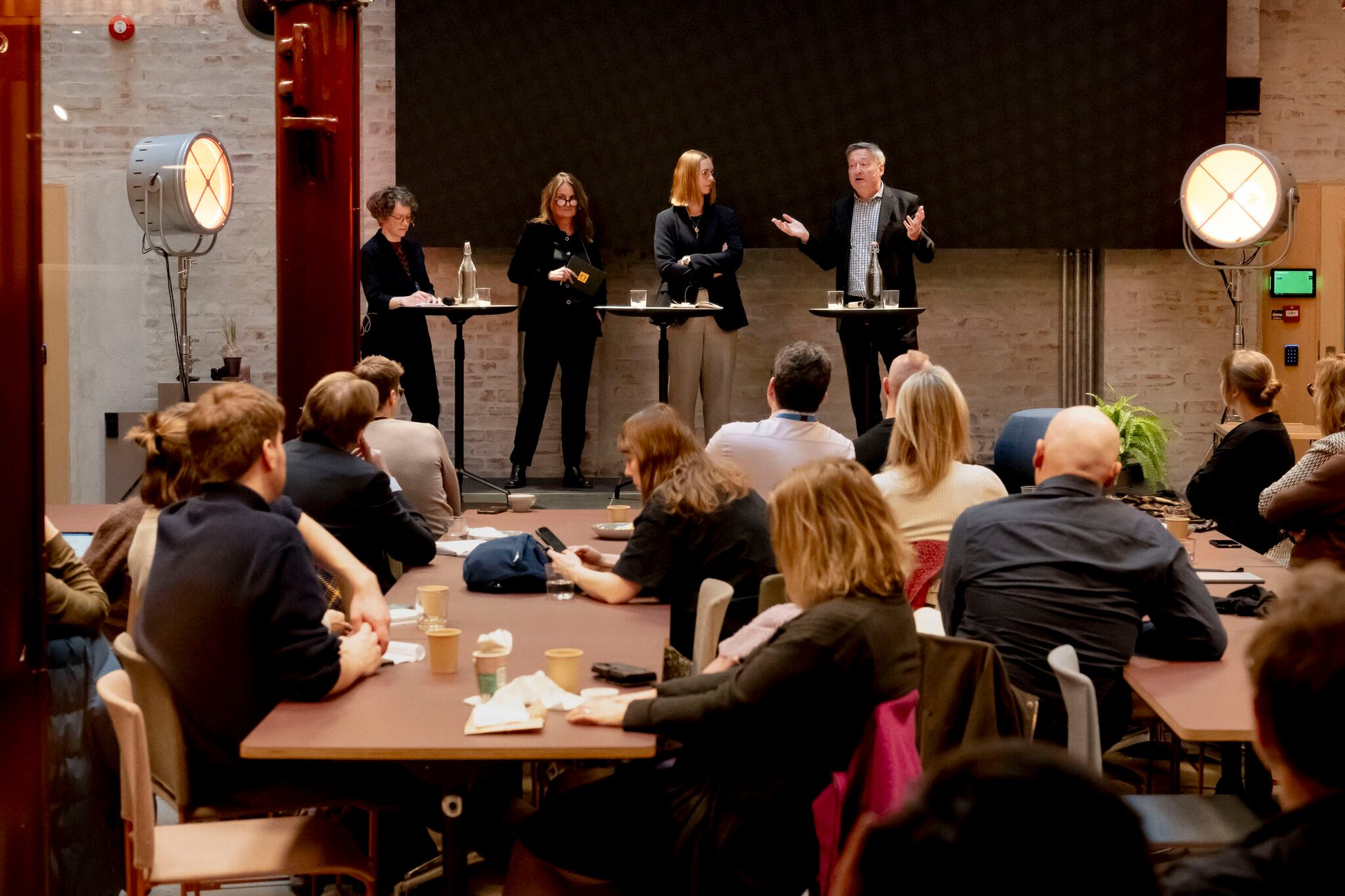 A panel of four speakers addresses an audience seated at tables in a dimly lit industrial-style room.