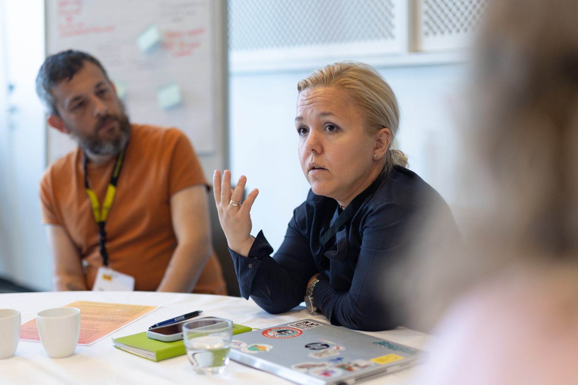 A woman gestures while speaking at a meeting, with a man blurred in the background.