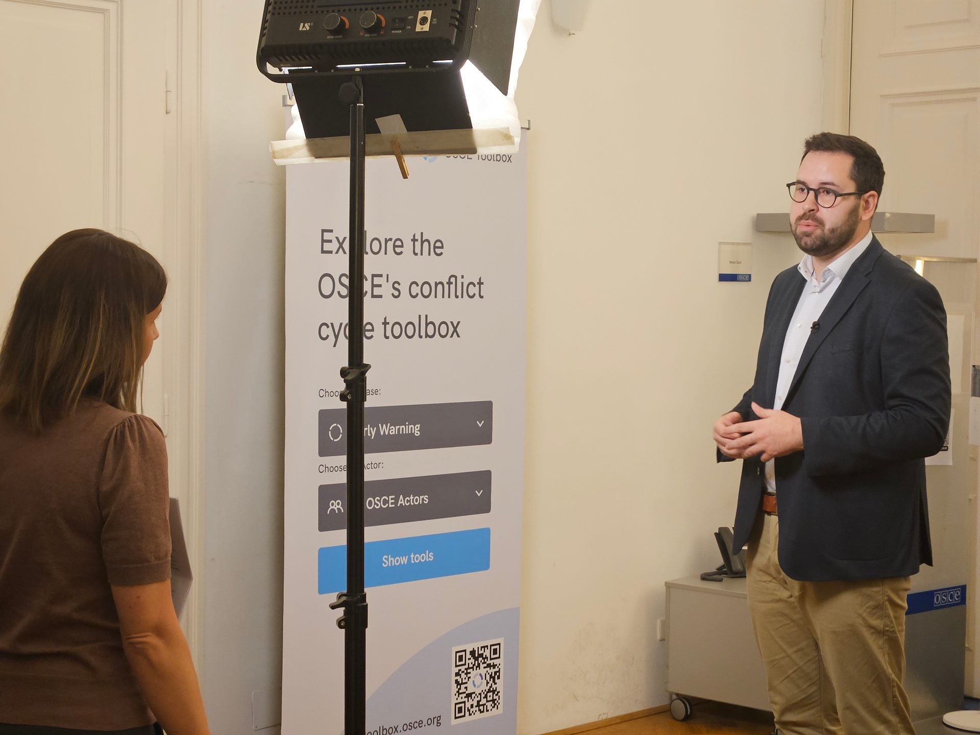 A man and woman next to a banner about the OSCE's conflict cycle toolbox, with a film light.