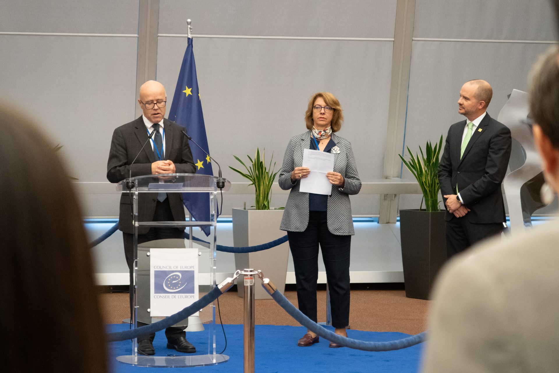 Three people, two men and one woman, stand at a podium during a Council of Europe event.