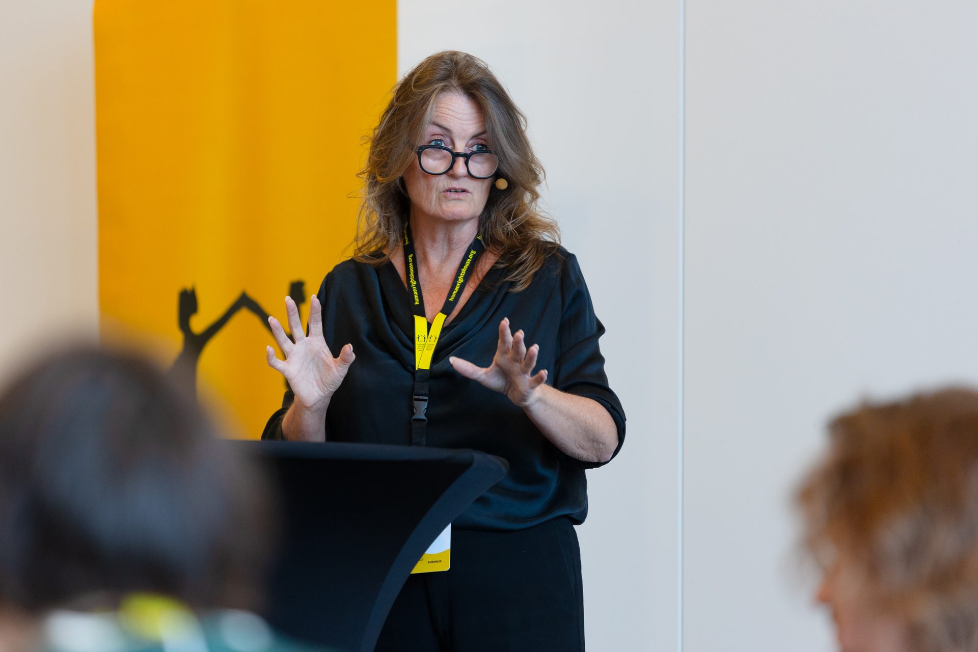 A woman with glasses and a black top speaks at a podium, gesturing with her hands, in front of a yellow banner.