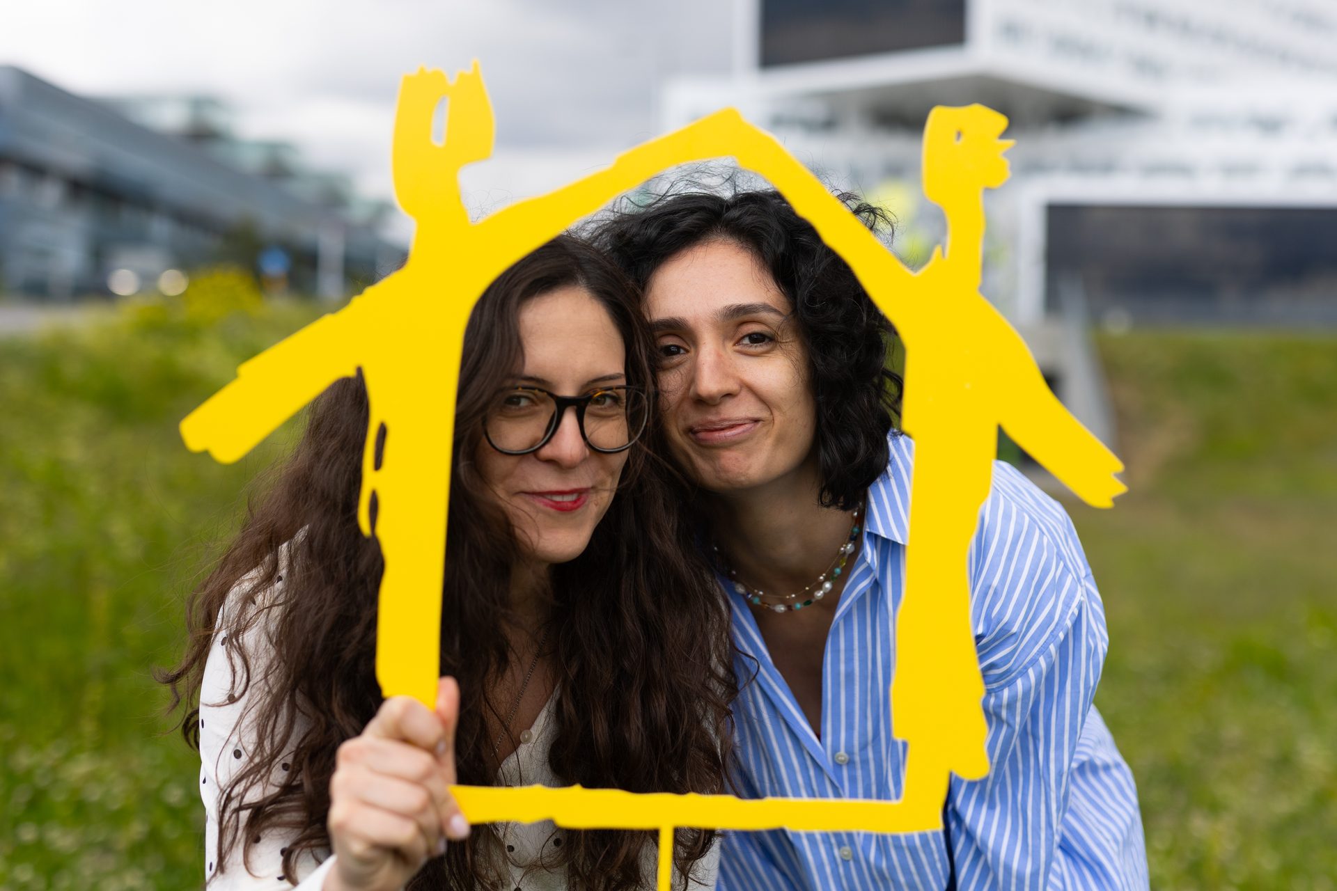 Two women smile through a yellow house frame, symbolizing home or real estate.