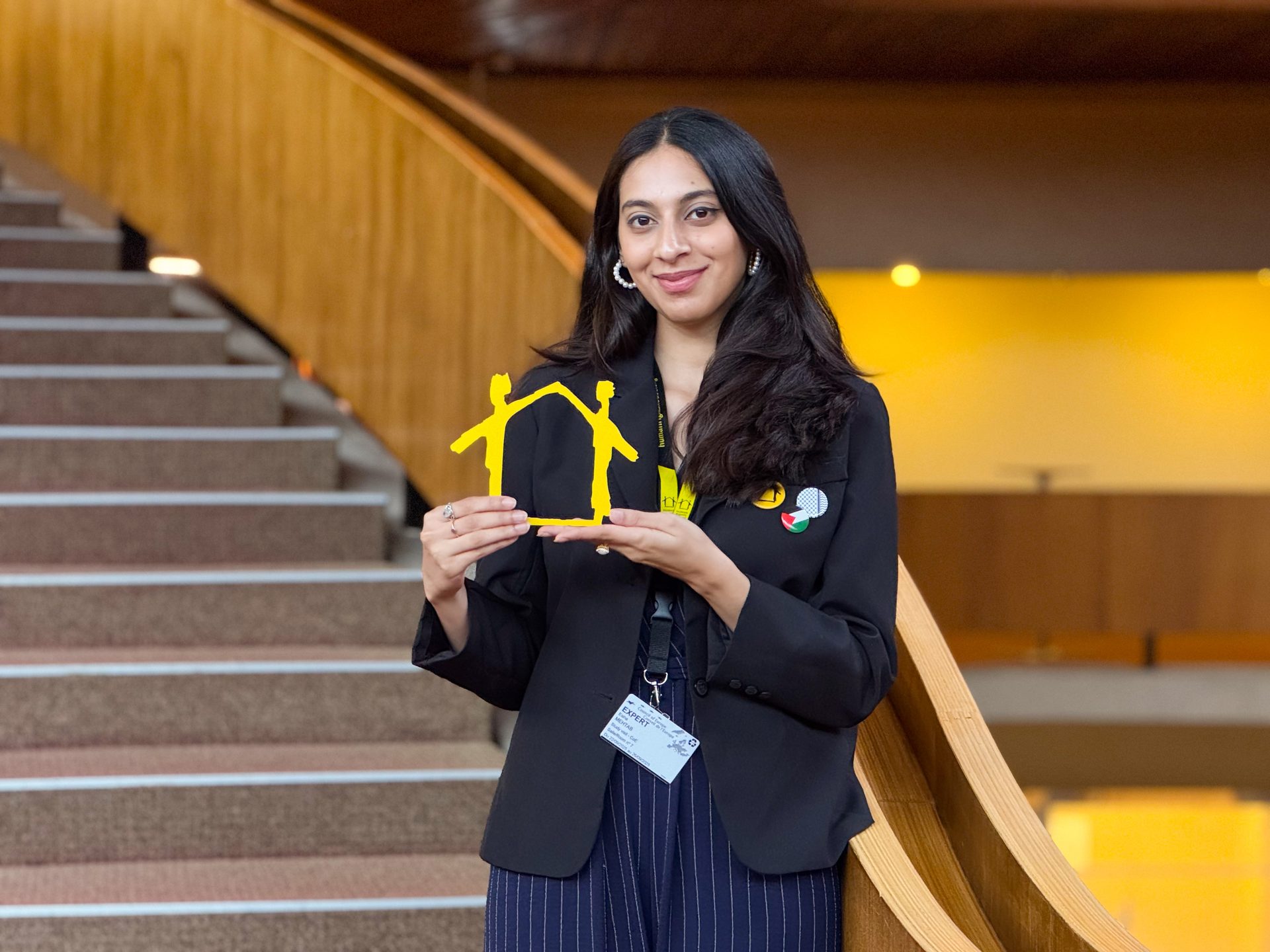 Young woman holding a yellow cutout of two figures forming a house, standing by a modern staircase.