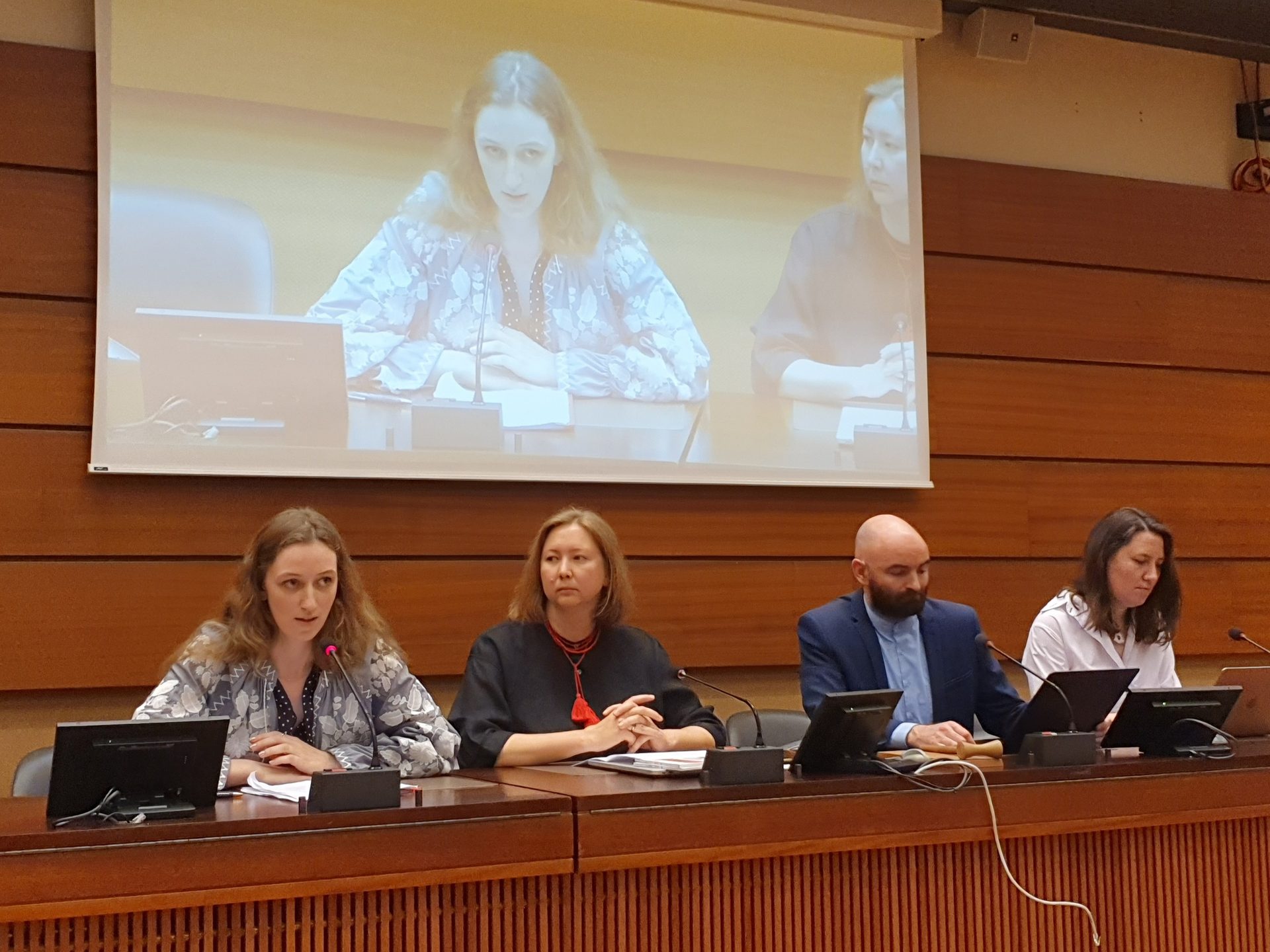 A panel of four people at a table with two participants projected on a large screen behind them.