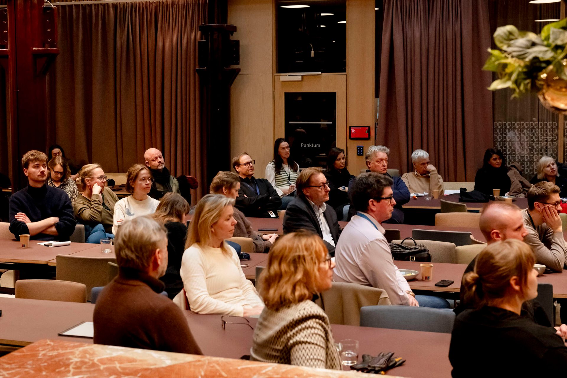 An audience of diverse individuals sits attentively at tables in a conference room, listening to a presentation.
