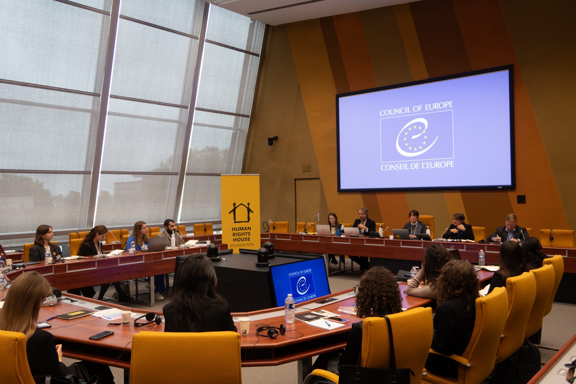 A U-shaped conference table with people attending a meeting, a large screen shows "COUNCIL OF EUROPE".